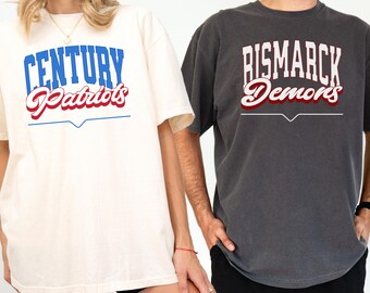 May include: Two t-shirts with vintage-style text graphics. The white t-shirt has blue and red text that reads "Century Patriots". The gray t-shirt has white and red text that reads "Bismarck Demons".