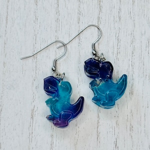 May include: A pair of blue and purple dinosaur earrings. The earrings are made of resin and have a silver hook.