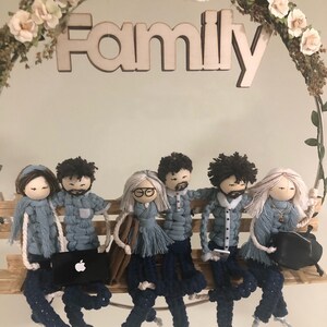May include: A wooden sign with the word "Family" hanging above six crocheted dolls sitting on a wooden bench. The dolls are wearing blue and white clothing and have different hairstyles.