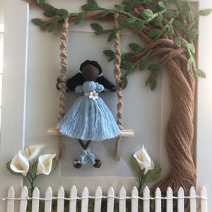 May include: A framed artwork featuring a brown yarn tree with green paper leaves. A doll with brown skin and blue yarn dress is on a swing made of brown yarn. White paper flowers and a white picket fence are at the bottom of the frame.