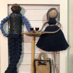 Op de afbeelding: Een ingelijst kunstwerk met twee figuren gemaakt van garen en stof. De figuren zijn een man en een vrouw, beiden gekleed in blauw. De man draagt een zwart vest en een bril. De vrouw draagt een blauwe jurk en heeft grijs haar. Ze staan naast een kleine tafel met twee glazen wijn erop. De vrouw houdt een kleine bruine tas vast met het woord "Vogue" erop.