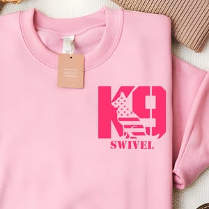 May include: A light pink sweatshirt with a crew neck. The sweatshirt features a magenta graphic of a dog silhouette within the letters "K9" and the word "SWIVEL". A blank tag is attached to the neckline.