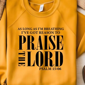 May include: Mustard yellow sweatshirt with the text "AS LONG AS I'M BREATHING I'VE GOT REASON TO PRAISE THE LORD" in black. The text "PSALM 15:06" is below the main text. The sweatshirt is folded on a neutral surface.