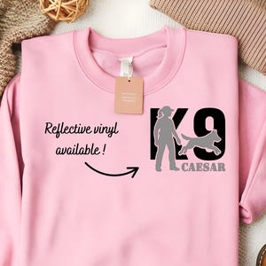 May include: A light pink sweatshirt with the text "K9 CAESAR" and a graphic of a person and dog. The sweatshirt also has the text "Reflective vinyl available!" with an arrow pointing to the graphic.