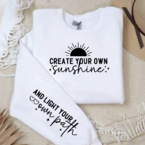 May include: White sweatshirt with black text that reads "Create your own sunshine" and a sun graphic. The sleeve has black text that reads "And light your own path" with two hearts.