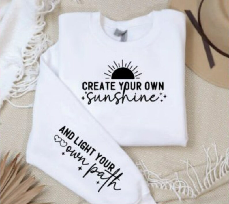 May include: White sweatshirt with black text that reads "Create your own sunshine" and a sun graphic. The sleeve has black text that reads "And light your own path" with two hearts.