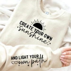 May include: A cream colored sweatshirt with the text "Create your own sunshine" and a black sun graphic on the front. The sleeve has the text "And light your own path" with two small hearts.