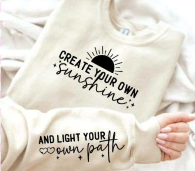 May include: A cream colored sweatshirt with the text "Create your own sunshine" and a black sun graphic on the front. The sleeve has the text "And light your own path" with two small hearts.