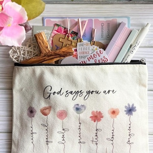 Peut inclure: Trousse en toile crème avec fermeture éclair noire, imprimée des mots "Dieu dit que tu es" et de motifs floraux. Contient des stylos, des autocollants et une croix en bois. Des versets bibliques sont imprimés en bas.