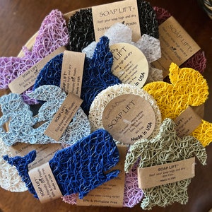 May include: A collection of colorful soap lifts in various shapes, including whales and turtles, displayed in a wooden bowl. The lifts are made of eco-friendly plastic and feature attached tags with product information. The text "SOAP LIFT" is visible on the tags.