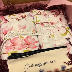 May include: Two rectangular packages wrapped in white paper with a pink floral magnolia pattern, tied with white ribbon featuring small gold crosses. A cream-colored canvas pencil case with a black zipper and the text God says you are printed on it.