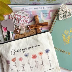 May include: A canvas zipper pouch with the text God says you are and floral illustrations, accompanied by a green notebook, a wooden cross, a white woven heart, a sticker reading pray more worry less, and various stationery items including pens.