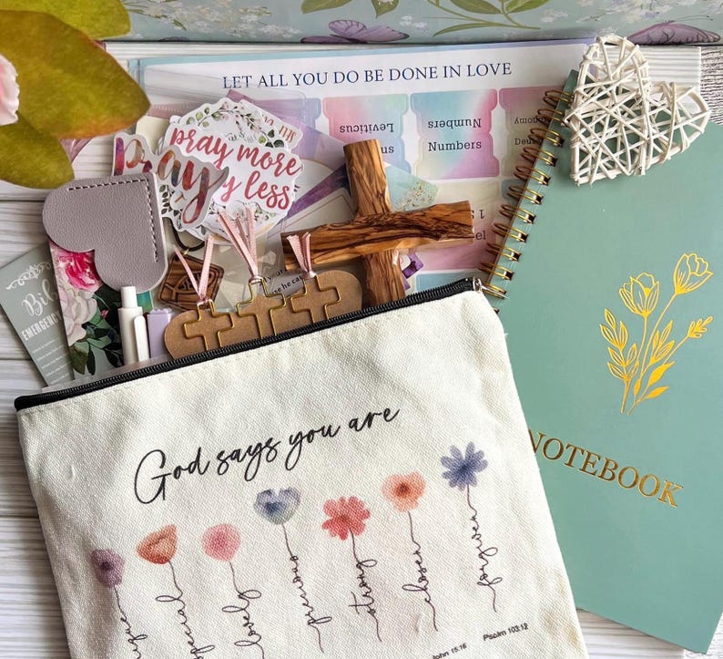 May include: A canvas zipper pouch with the text God says you are and floral illustrations, accompanied by a green notebook, a wooden cross, a white woven heart, a sticker reading pray more worry less, and various stationery items including pens.