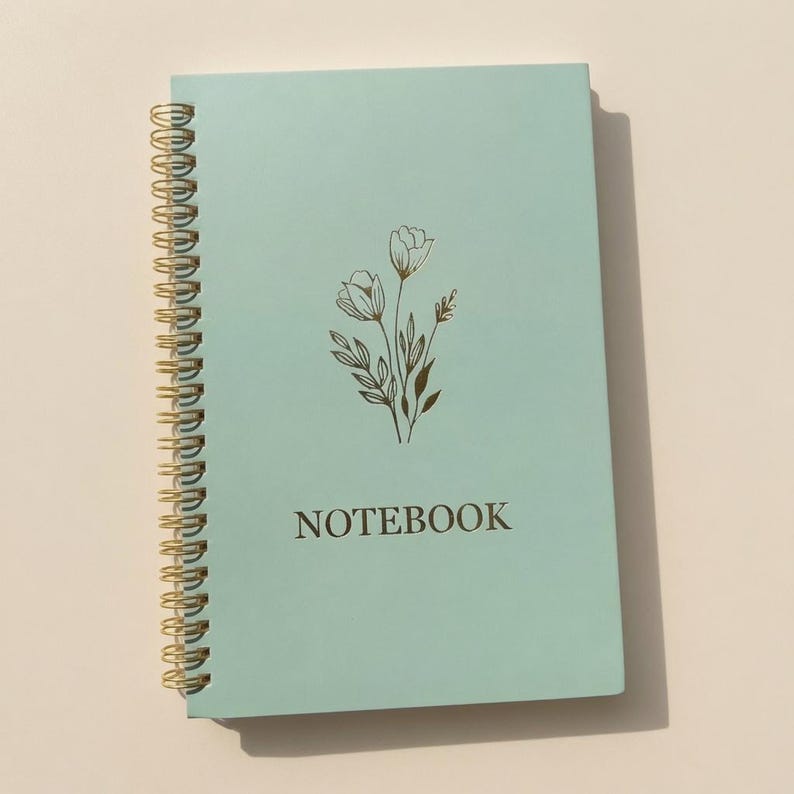 May include: A light mint green spiral-bound notebook featuring a gold-colored line art illustration of a floral bouquet centered on the front cover. The word NOTEBOOK is printed in gold capital letters below the floral design on the cover.
