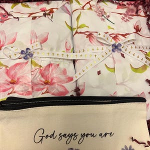 May include: A beige canvas pencil case with the black text God says you are and a floral print at the bottom, placed in front of two white packages wrapped in floral paper, tied with white ribbon featuring gold crosses and purple flower accents.