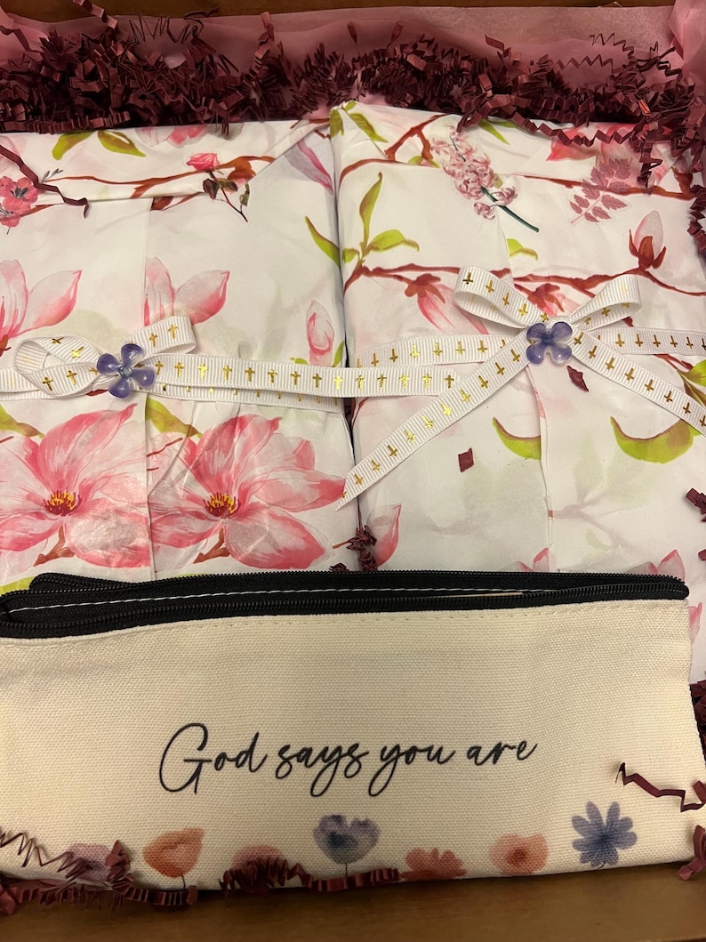 May include: A beige canvas pencil case with the black text God says you are and a floral print at the bottom, placed in front of two white packages wrapped in floral paper, tied with white ribbon featuring gold crosses and purple flower accents.