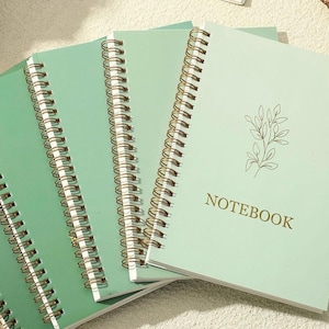 May include: A set of three spiral-bound notebooks with light green covers. Each notebook features a gold-colored wire coil binding and a delicate line-art illustration of a leafy branch. The word NOTEBOOK is printed in gold capital letters.