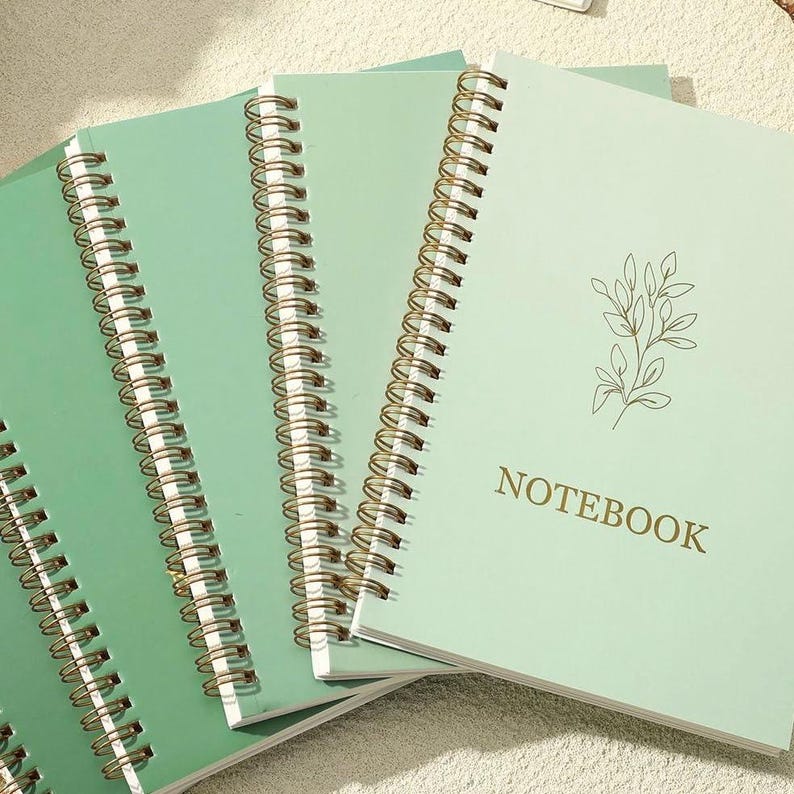 May include: A set of three spiral-bound notebooks with light green covers. Each notebook features a gold-colored wire coil binding and a delicate line-art illustration of a leafy branch. The word NOTEBOOK is printed in gold capital letters.