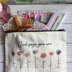 May include: A cream-colored canvas zipper pouch featuring the text God says you are above seven watercolor-style flowers with stems labeled unique, special, lovely, precious, strong, chosen, and forgiven, with Bible verse references printed below.