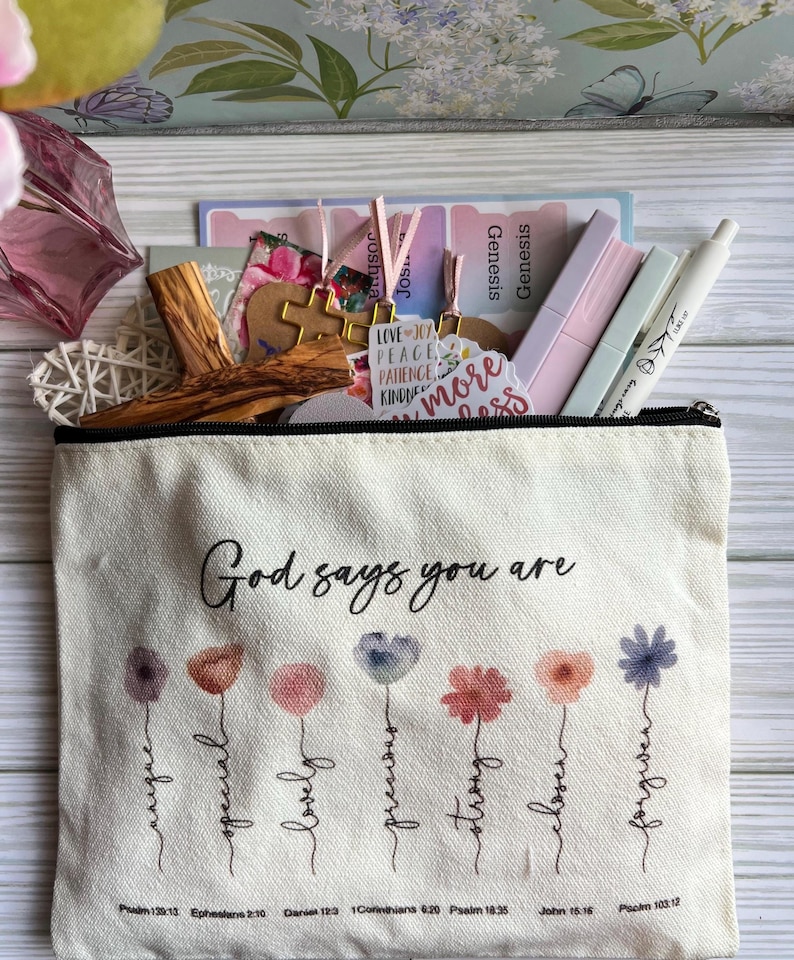 May include: A cream-colored canvas zipper pouch featuring the text God says you are above seven watercolor-style flowers with stems labeled unique, special, lovely, precious, strong, chosen, and forgiven, with Bible verse references printed below.