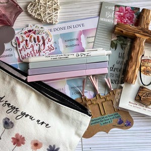 May include: A collection of religious stationery and accessories including a canvas pouch with floral print, a wooden cross, a wooden Jesus pendant, four pastel pens, a sticker reading Pray, and a card with the text Beloved, let us love one another.