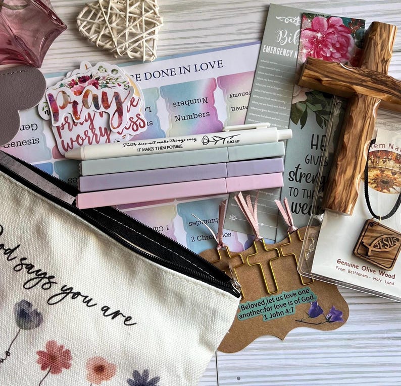 May include: A collection of religious stationery and accessories including a canvas pouch with floral print, a wooden cross, a wooden Jesus pendant, four pastel pens, a sticker reading Pray, and a card with the text Beloved, let us love one another.