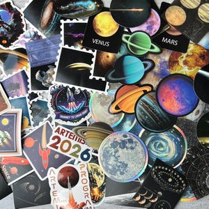 Op de afbeelding: Een verzameling ruimtevaartstickers met planeten, raketten en hemellichamen. Zichtbare tekst bevat VENUS, MARS, JOURNEY TO LUNAR ORBIT en ARTEMIS 2026 PROGRAM. De stickers tonen diverse kleuren, waaronder blauw, oranje en zwart.