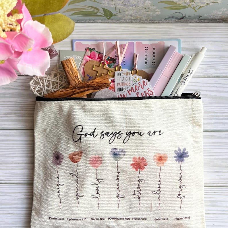 May include: A cream-colored canvas pouch featuring the text God says you are above seven watercolor-style flower illustrations. Each flower stem is labeled with words like unique, special, lovely, precious, strong, chosen, and forgiven, with Bible verses printed below.