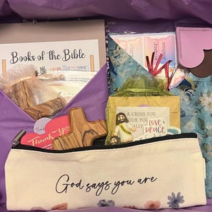May include: A gift box containing a spiral-bound Books of the Bible notebook, a floral-patterned pouch with heart-shaped tabs, a wooden cross, a small Jesus figurine, and a cream-colored pencil case with the text God says you are and floral accents.