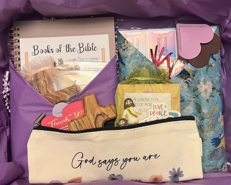 May include: A gift box containing a spiral-bound Books of the Bible notebook, a floral-patterned pouch with heart-shaped tabs, a wooden cross, a small Jesus figurine, and a cream-colored pencil case with the text God says you are and floral accents.