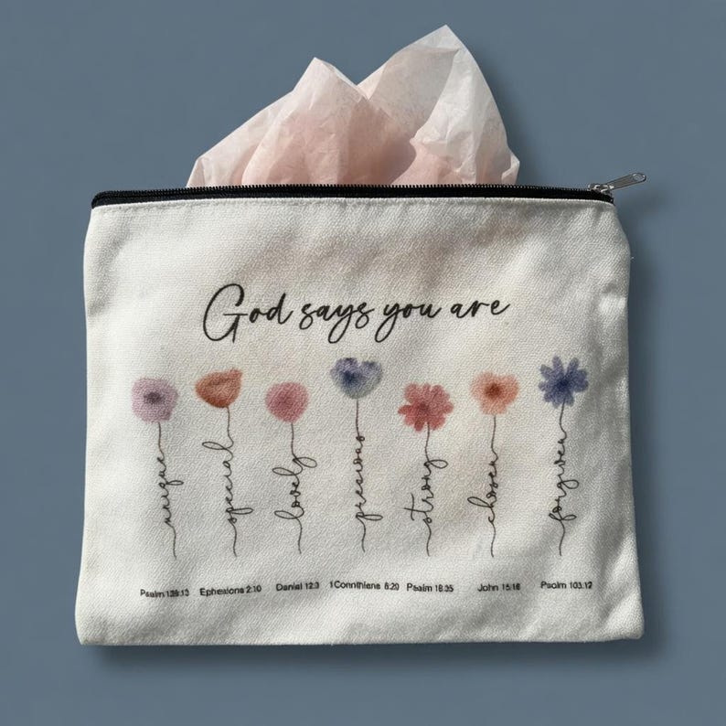 May include: A cream-colored canvas zipper pouch featuring the text God says you are above seven watercolor-style flower illustrations. Each flower stem is formed by a cursive word and includes a small Bible verse reference printed below it.