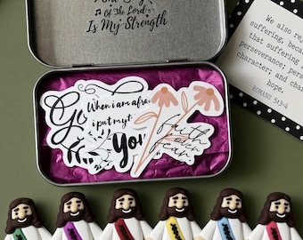 May include: A rectangular metal tin with a hinged lid featuring the text The Joy of the Lord Is My Strength. Inside are stickers on purple tissue paper and six small white figurines of a bearded figure wearing colored sashes with text.