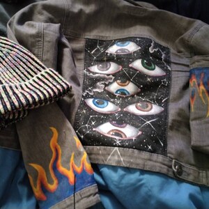 May include: A gray denim jacket with a black patch on the back featuring multiple colorful eyes and a starry sky design. The jacket has orange and yellow flames painted on the sleeves.