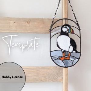 May include: Stained glass art piece featuring a puffin bird. The bird has a white chest, black back, and colorful beak. The artwork is framed in black metal and hangs from a chain. The word "Template" is written in white script.