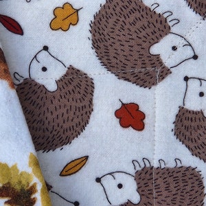 May include: White fabric with a repeating pattern of brown hedgehogs and orange and yellow leaves. The hedgehogs are facing different directions and have black eyes.