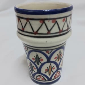 May include: A white ceramic cup with blue and red geometric patterns. The cup has a wide base and a narrow top.