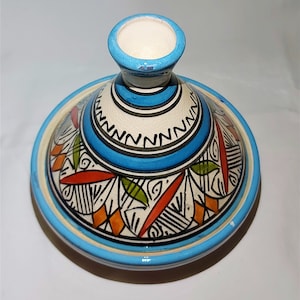 May include: A blue and white ceramic tagine lid with a geometric pattern in black, orange, and green. The lid has a raised center with a blue rim.