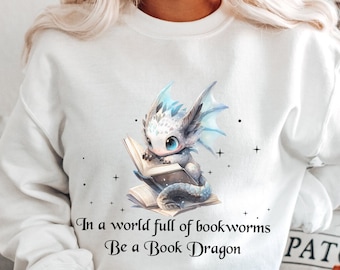 Può includere: Felpa bianca con un drago blu e bianco che legge un libro. Il testo sulla felpa recita "In un mondo pieno di vermi dei libri Sii un drago dei libri".
