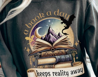 Puede incluir: Una sudadera gris oscuro con un diseño gráfico de una pila de libros, una luna creciente, una montaña y un dragón. El texto "a book a day keeps reality away" está impreso en la sudadera.