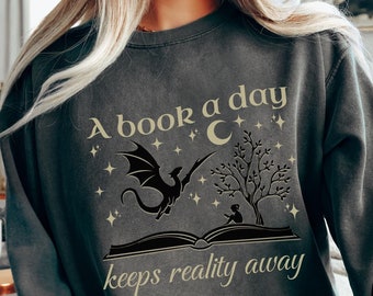 Puede incluir: Una sudadera gris oscuro con un estampado gráfico de un dragón volando sobre un libro con una luna creciente y estrellas. El texto "A book a day keeps reality away" está impreso debajo del gráfico.