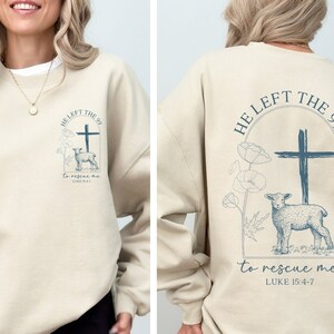 Pu&ograve; includere: Una felpa beige con una stampa grafica blu sul davanti e sul retro. La grafica presenta una croce, un agnello e il testo "HE LEFT THE 99 to rescue me LUKE 15:4-7".