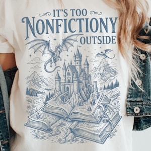 May include: An ivory t-shirt featuring a blue graphic print of a castle emerging from open books, surrounded by dragons and mountains. The text reads IT'S TOO NONFICTIONY OUTSIDE. The shirt is layered under a blue denim jacket.