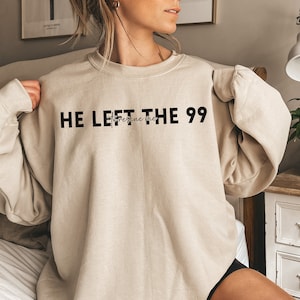 May include: A beige sweatshirt with the text "HE LEFT THE 99" in black block letters. Below the text is the phrase "to rescue me" in a cursive font. The sweatshirt is a neutral color and has long sleeves.
