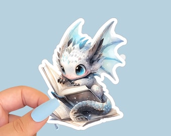 May include: A sticker depicting a cartoon dragon with white and blue scales, big blue eyes, reading a book. The sticker is held by a hand with blue nails. The background is a light blue. The dragon is the central focus of the image.
