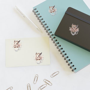 May include: A light blue and white striped notebook with a black notebook on top. Both notebooks have stickers of a small, brown dragon in a teacup. The notebooks are on a white surface with silver paper clips.