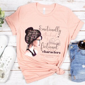 May include: A peach-colored t-shirt with a graphic of a woman with a stack of books for hair, wearing glasses and reading a book. The text on the shirt reads "Emotionally attached to fictional characters *minor emotional damage may occur*"