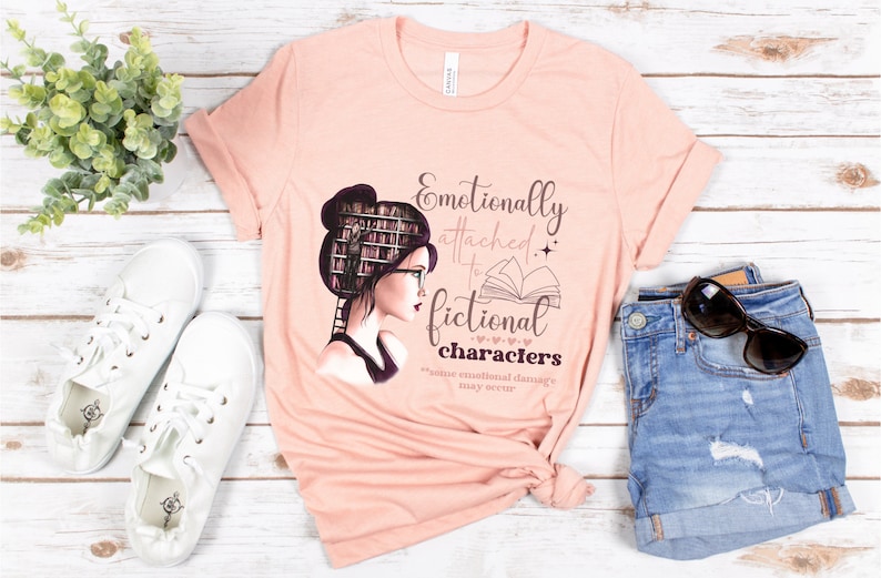 May include: A peach-colored t-shirt with a graphic of a woman with a stack of books for hair, wearing glasses and reading a book. The text on the shirt reads "Emotionally attached to fictional characters *minor emotional damage may occur*"