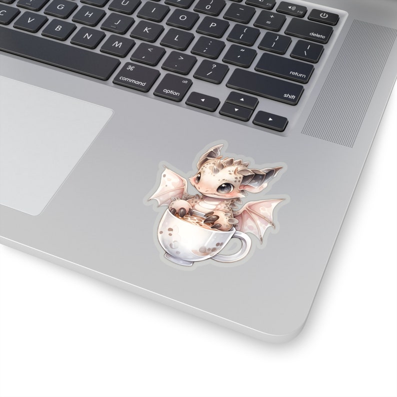 May include: A small, white dragon with brown spots sits in a white mug with a brown liquid. The dragon has large, brown eyes and pink wings.