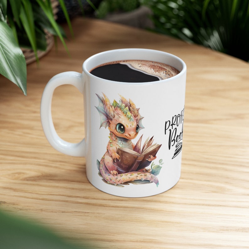 Op de afbeelding: Witte keramische mok met een kleurrijke illustratie van een draak die een boek leest. De tekst "Professional Bookworm" staat op de mok gedrukt.