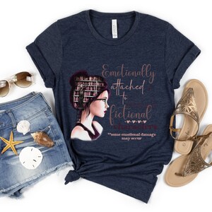 May include: A navy blue t-shirt with a graphic of a woman with books stacked on her head. The text on the shirt reads "Emotionally attached to fictional characters" with the words "some emotional damage may occur" below.